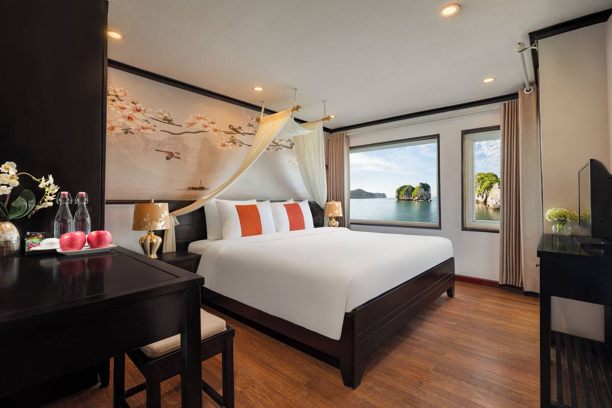 A hotel room with a bed, a desk, a chair, a TV, a window with a view of a body of water and a mountain, and a wall with a floral mural.