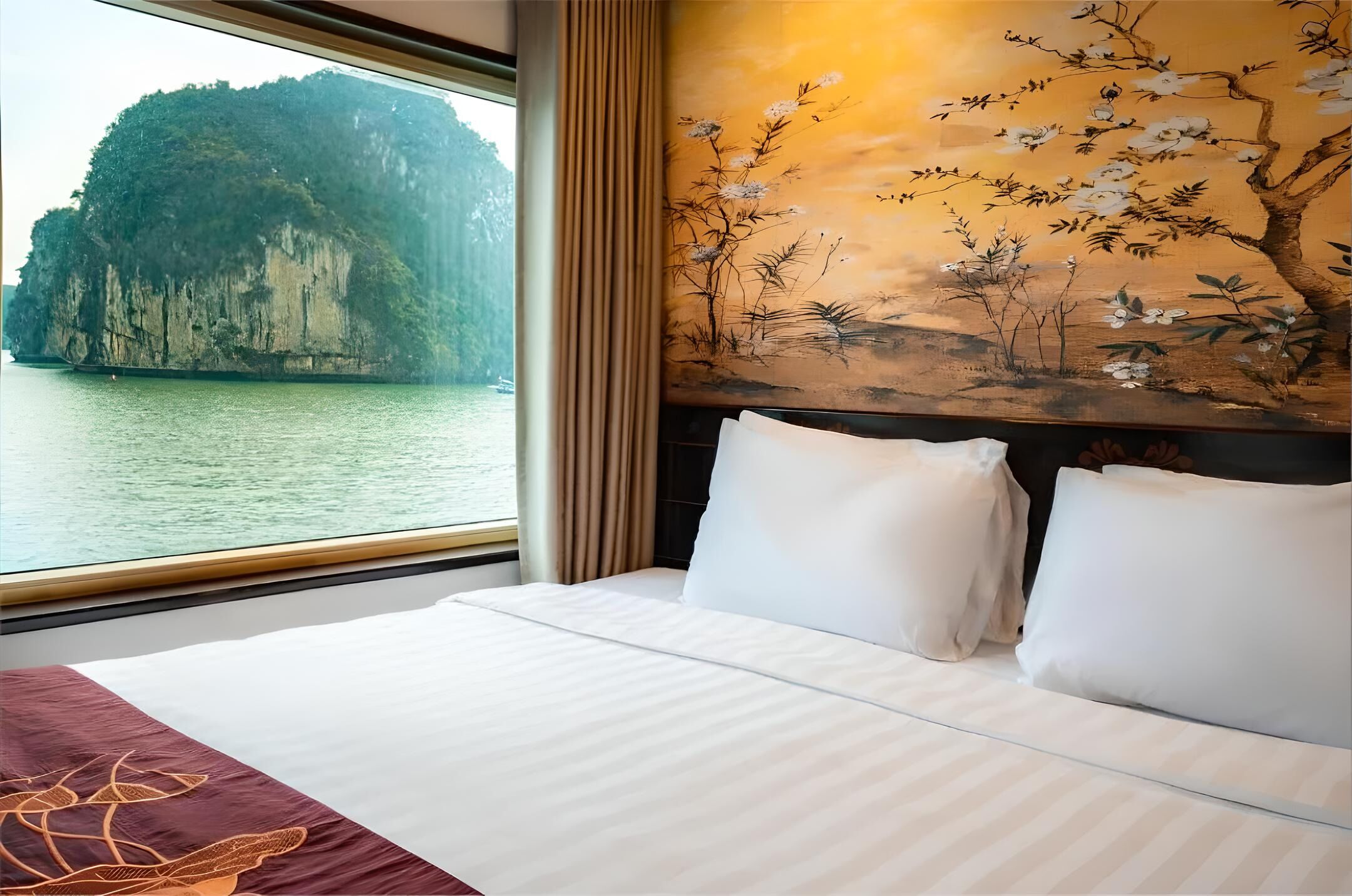 A hotel room with a large bed, a view of a mountainous island through the window, and a painting of flowers and trees on the wall.