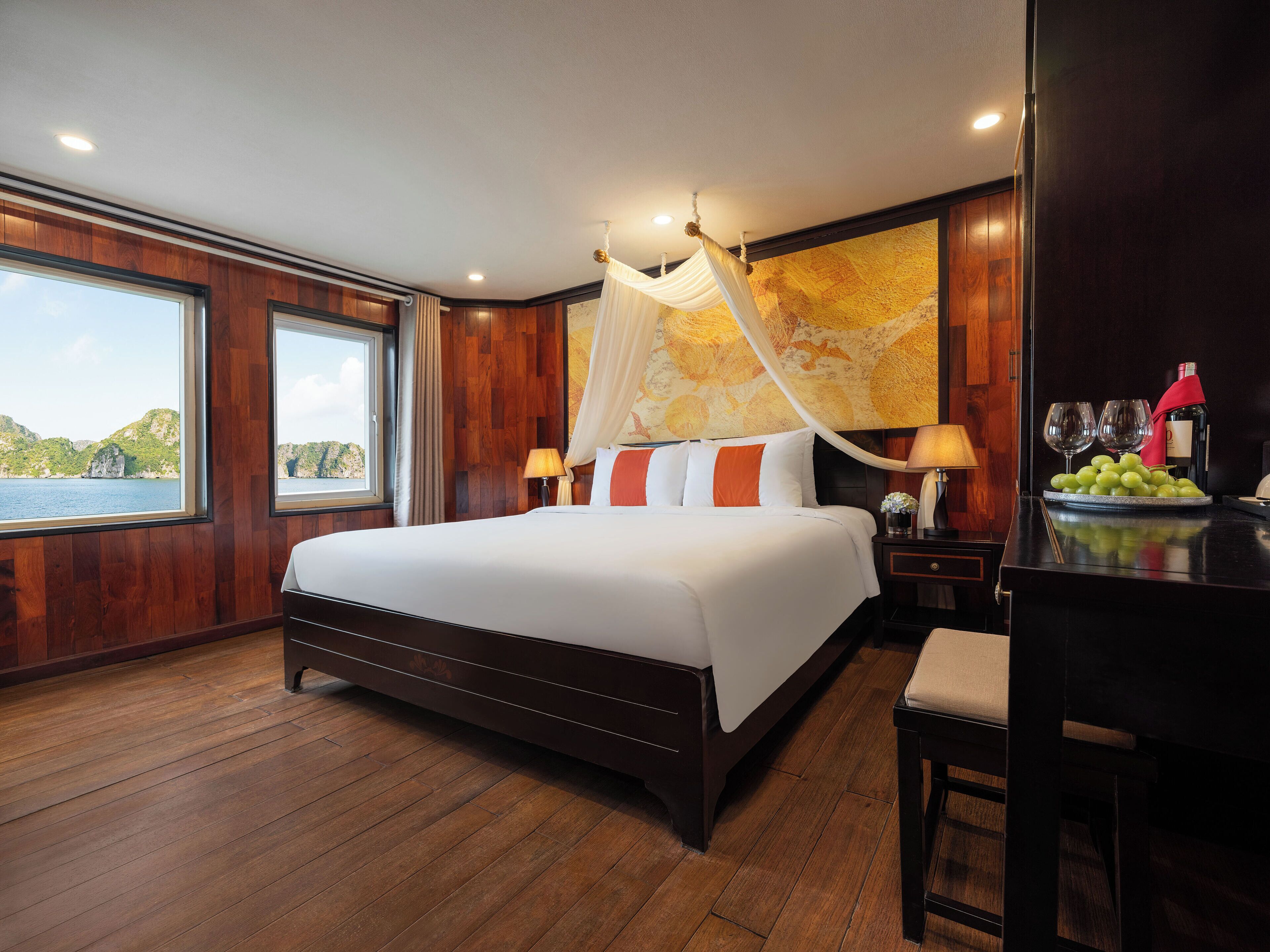 A large bed with a canopy, a nightstand with a wine bottle and glasses, a fruit bowl, and a view of the sea through a window.