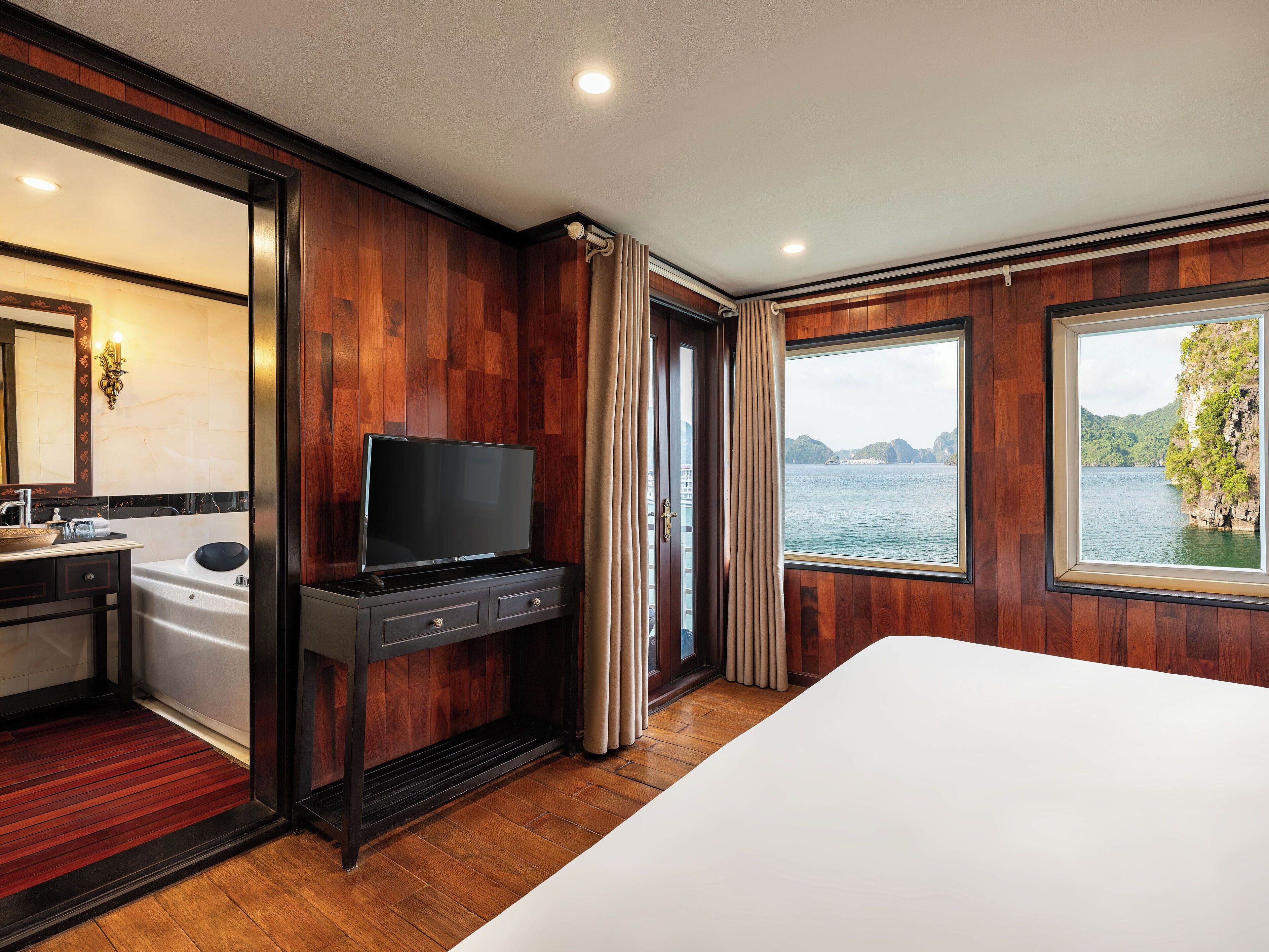 A hotel room with a wooden interior, a large bed, a TV, a bathtub, and a window with a view of a lake and mountains.