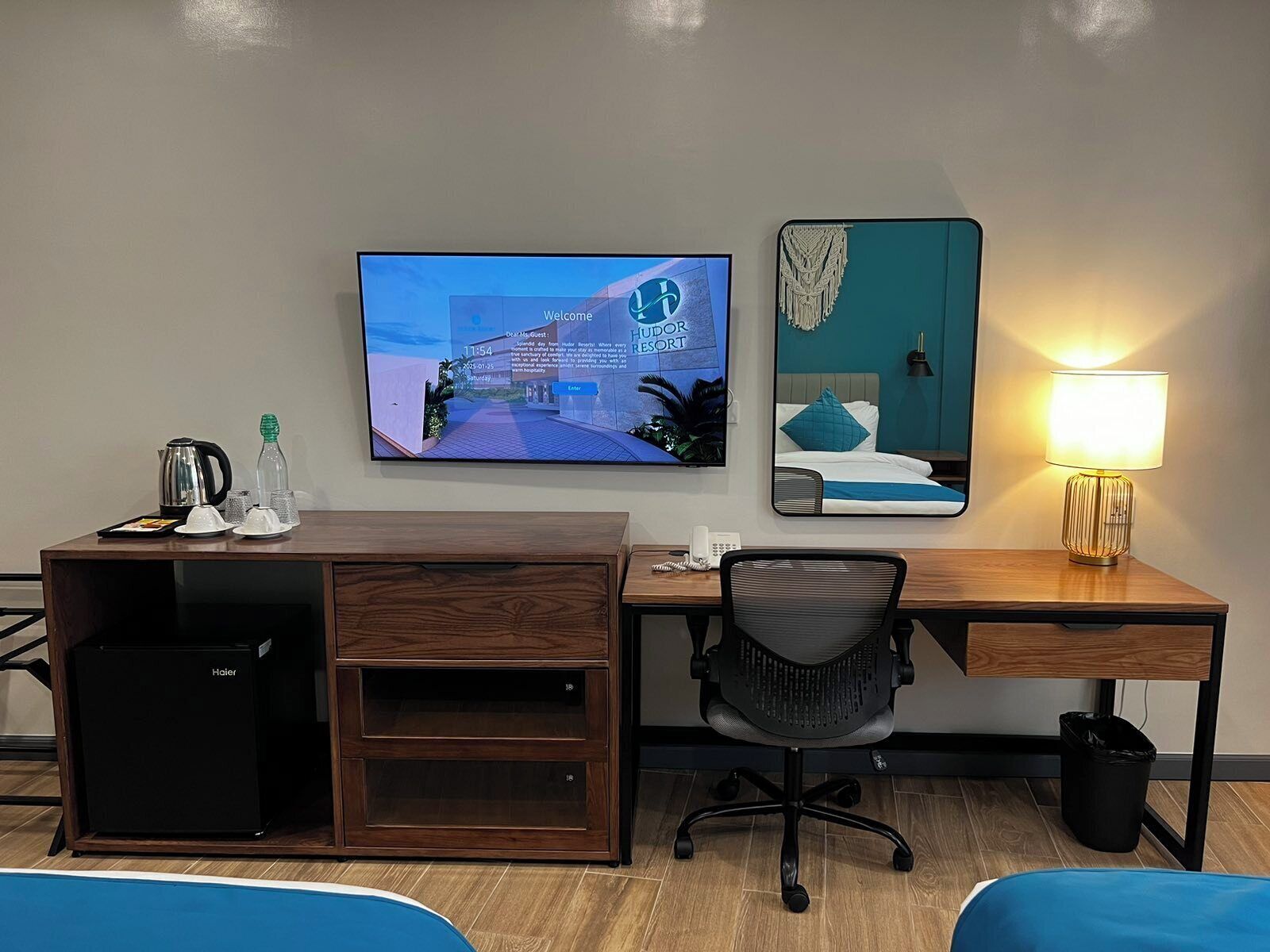 Ett hotellrum med ett skrivbord i trä, en tv som visar bilder från ett semesterparadis, en säng med en blå kudde och en sänglampa.