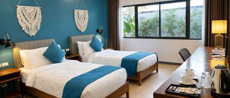 A hotel room with two beds, a desk with a lamp, a coffee maker, and a view of greenery outside.
