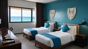 A hotel room with two beds, a desk, a view of the ocean, and macrame wall hangings. - Hudor Resort (Bauang)