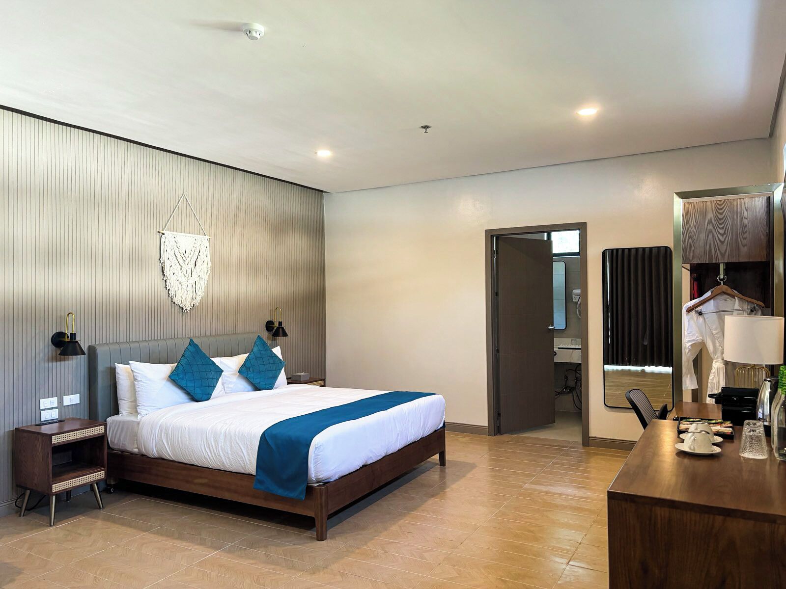 Una habitación de hotel moderna con una cama grande, lámparas de noche, un adorno de macramé en la pared y una mesita de noche de madera.