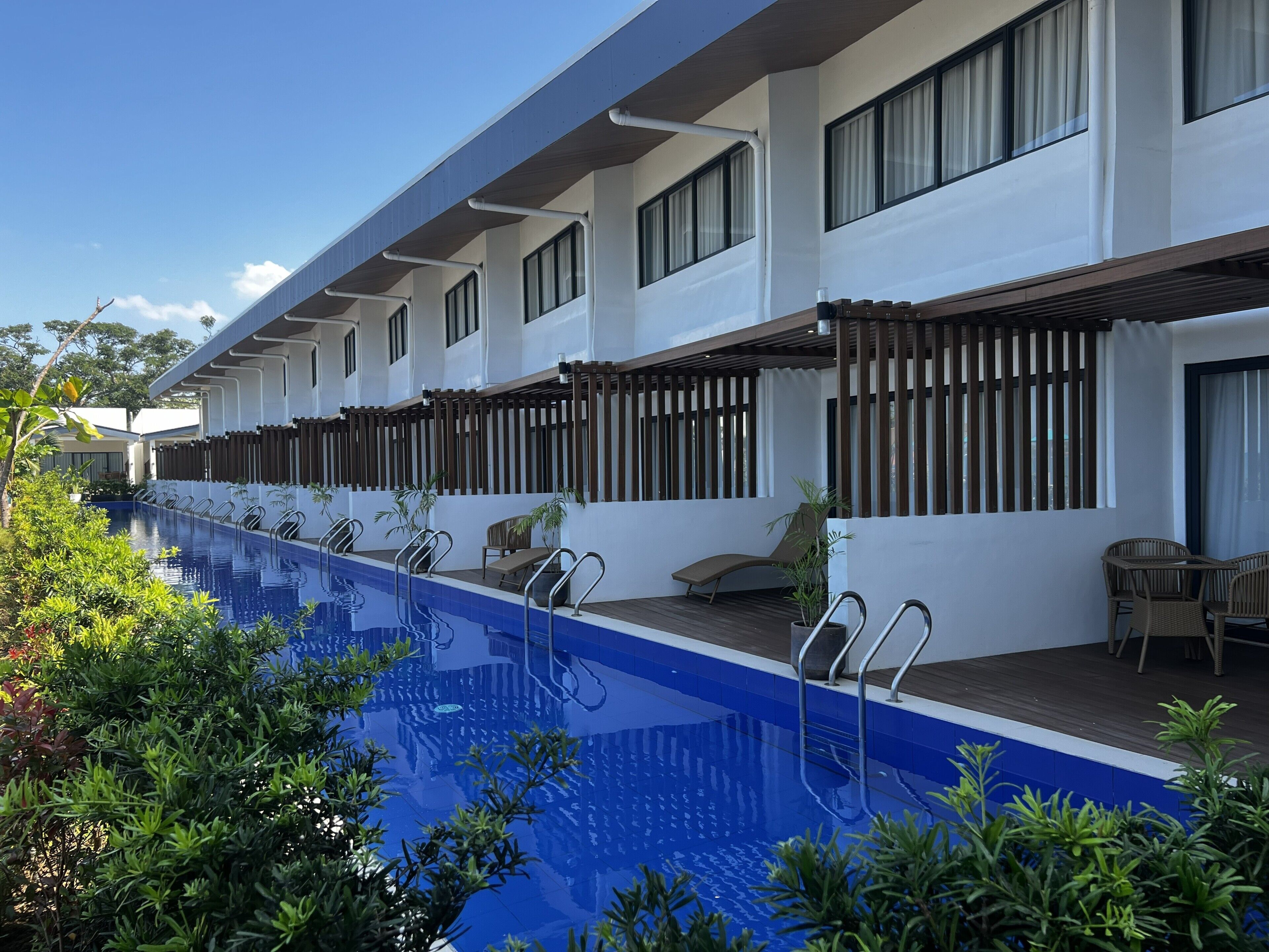 A hotel with a long swimming pool, outdoor seating, and a covered area with windows.