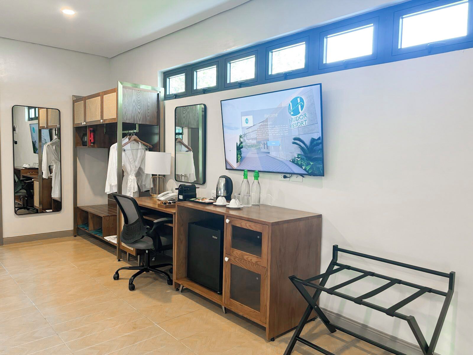 Habitación de hotel con escritorio, silla, armario y televisor mostrando el nombre del hotel.