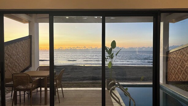 A balcony with a view of the ocean at sunset, a pool, and wicker furniture. - Hudor Resort (Bauang)