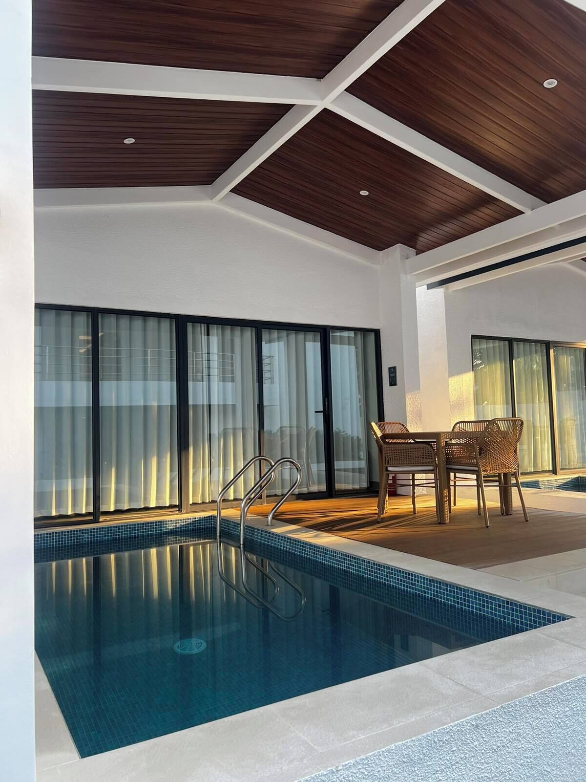 A modern indoor swimming pool with a clear view of the pool, a wooden ceiling, and a dining area with chairs and a table.