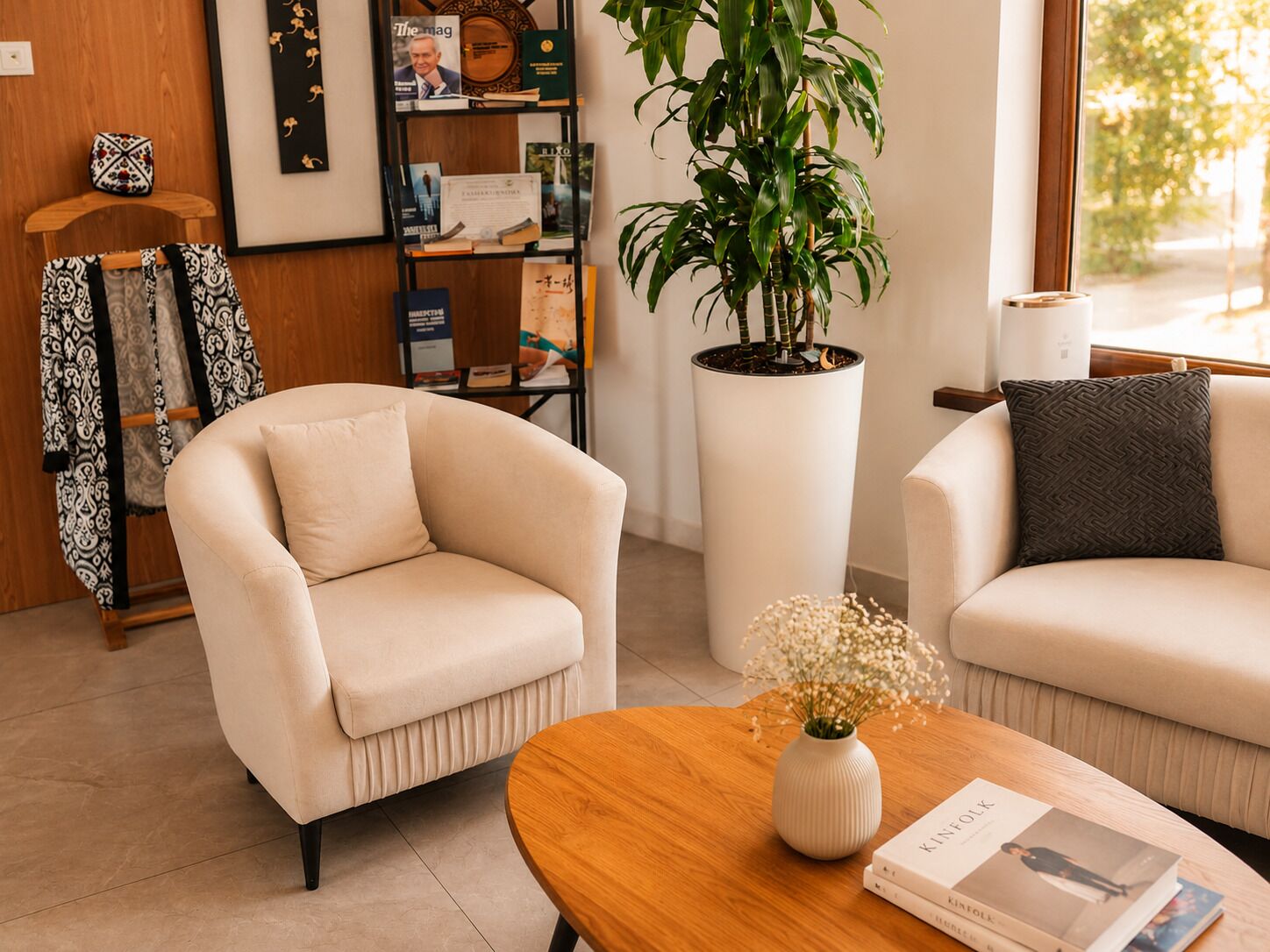 Un salon moderne avec deux fauteuils beiges, une table basse en bois, une plante en pot et une bibliothèque contenant divers objets.