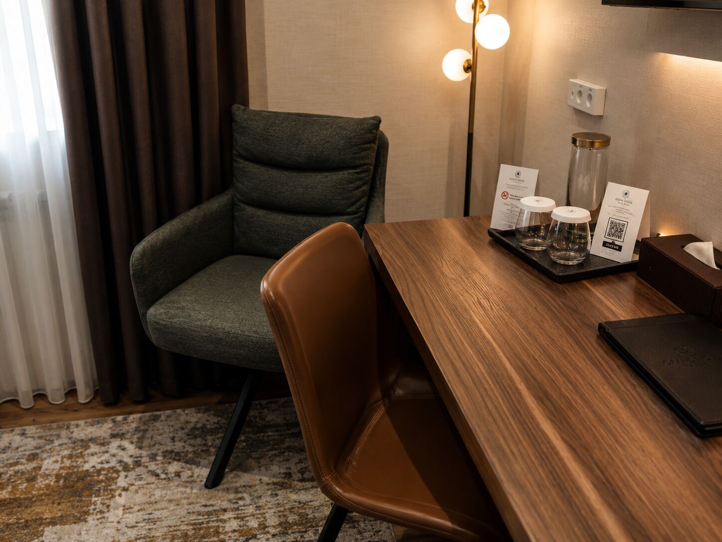 Habitación de hotel con una mesa de comedor de madera, una silla de cuero, un sillón verde oscuro, una lámpara de pie y una bandeja con vasos y un menú.