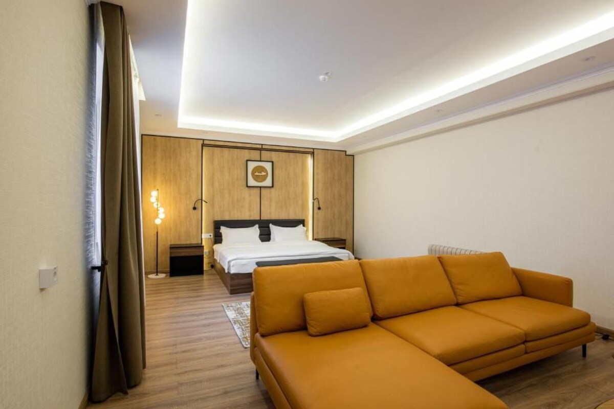A modern hotel room with a wooden floor, a large sofa, a bed with white linens, and a wooden wardrobe.