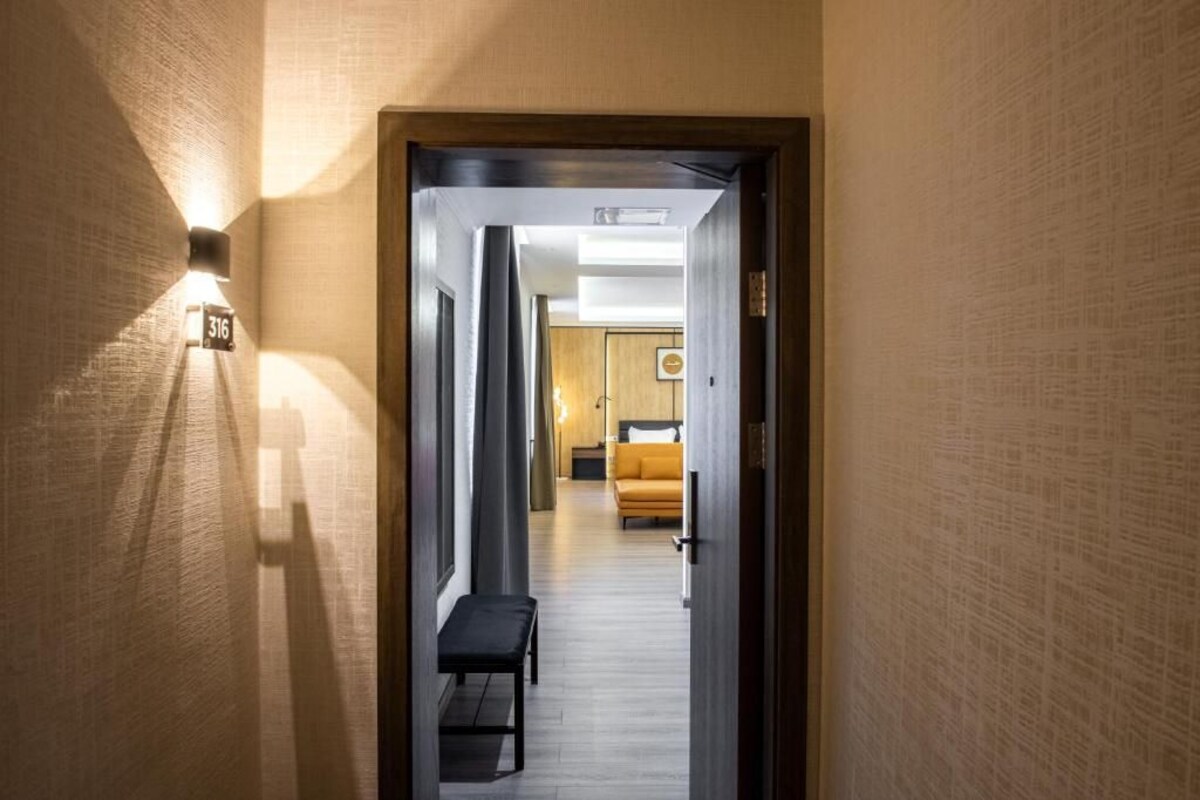 A hallway with a bench, a door with a number sign, and a room with a bed and a wall-mounted picture.