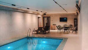 A modern indoor swimming pool with lounge chairs, a dining area, and a flat-screen TV mounted on the wall.