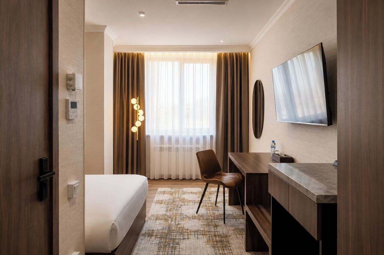 Habitación de hotel con cama, escritorio, silla, televisión y una ventana grande con cortinas.