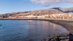 A coastal town with buildings along the beach, rocky shoreline, and mountains in the background. - Oasis Marítimo en Güímar (Puertito de Güímar)