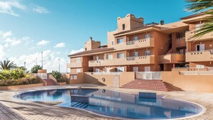 A swimming pool area with a tiled deck, a building with multiple balconies, and a clear blue sky. - Oasis Marítimo en Güímar (Puertito de Güímar)