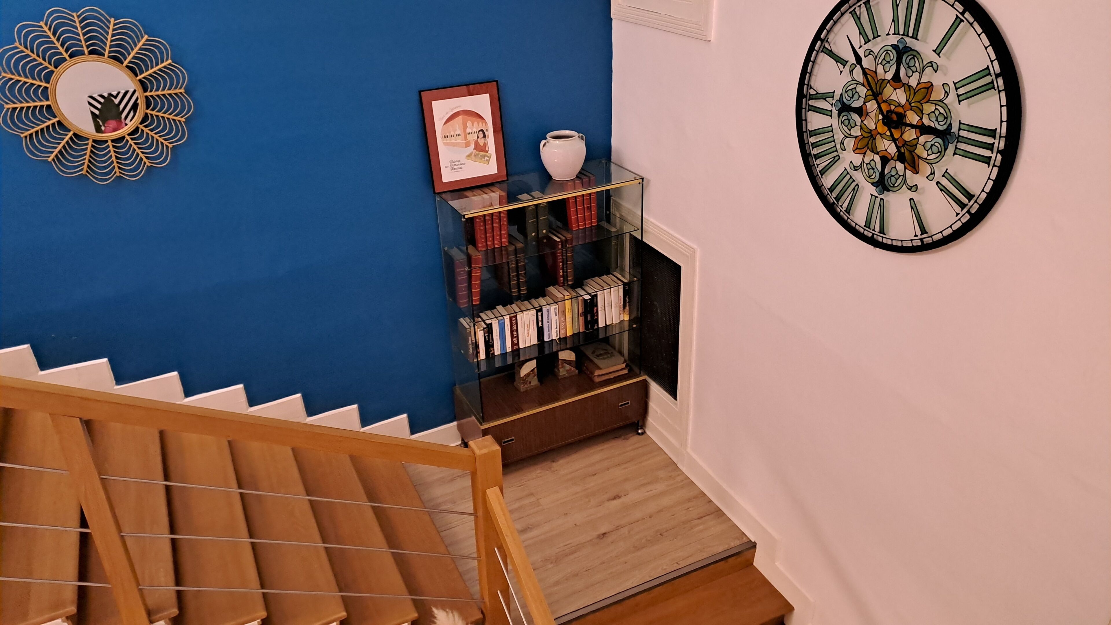 Un escalier avec une rampe en bois menant à un palier où se trouve une bibliothèque en verre adossée à un mur bleu.