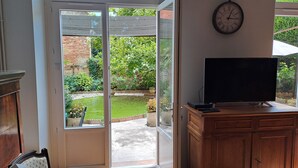 A room with a sliding glass door leading to a garden, a flat-screen TV on a wooden cabinet, and a wall clock. - Charmant Gite aux Beaux Volumes, Jardin Privé, Idéal en Familles (Beaumont-de-Lomagne)