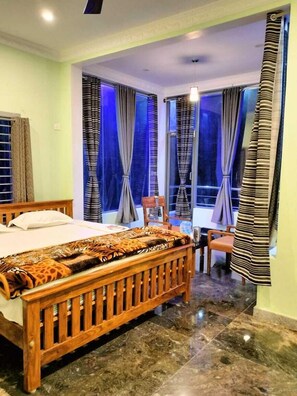 A bedroom with a wooden bed, a patterned bedspread, and a view of the outside through large windows with curtains. - Yelagiri Gold Hill Resort (Tirupattur)