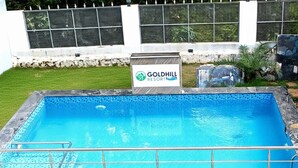 A rectangular swimming pool with a metal safety railing, surrounded by a stone border. A sign in the background reads "Goldhill Resort."