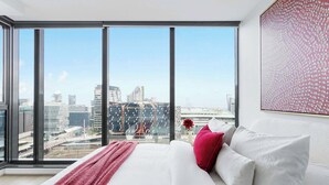 A modern hotel room with a large window offering a city view, a neatly made bed with white linens, and a red throw blanket. - A 2BR Apt with Harbour Views and FREE Parking (Melbourne)