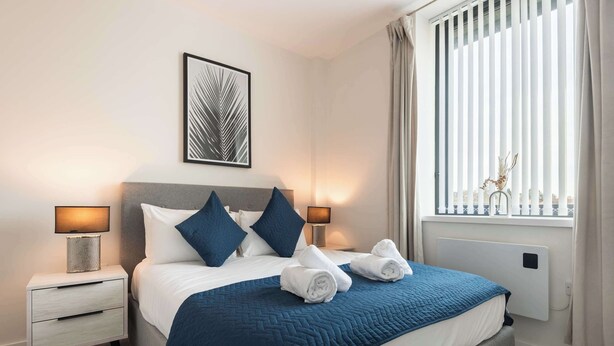 A neatly made bed with a blue quilt, two pillows, and two rolled towels on the side. - Modern & Cosy 1 Bedroom Apartment in York (York)