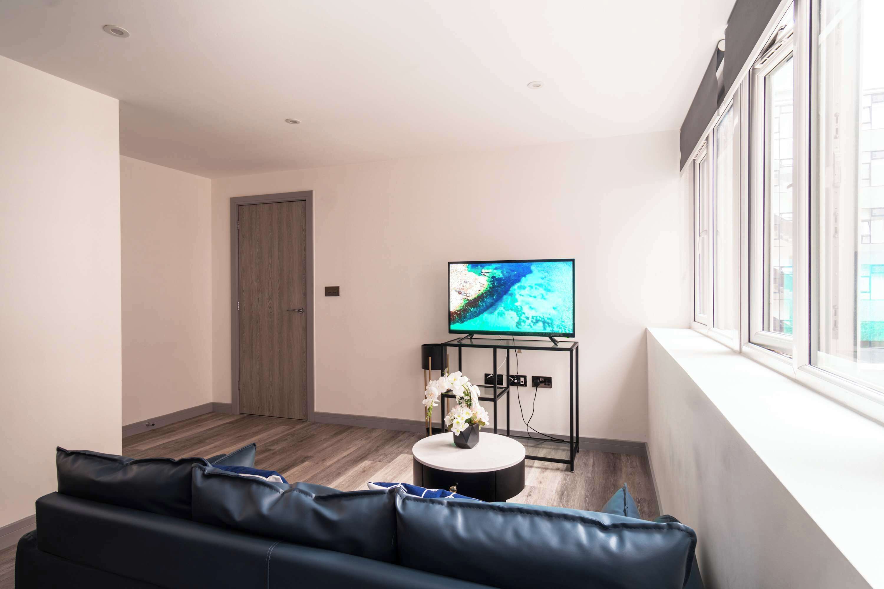 A modern living room with a dark leather sofa, a round coffee table with a vase of flowers, a flat-screen TV on a black stand, and a large window with a view of the outside.