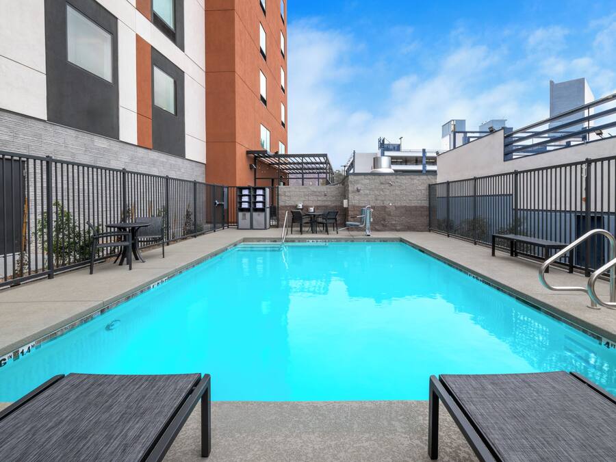 A rectangular swimming pool with a clear blue water surface, surrounded by a low brick wall, and adjacent to a modern building with large windows and a flat roof.