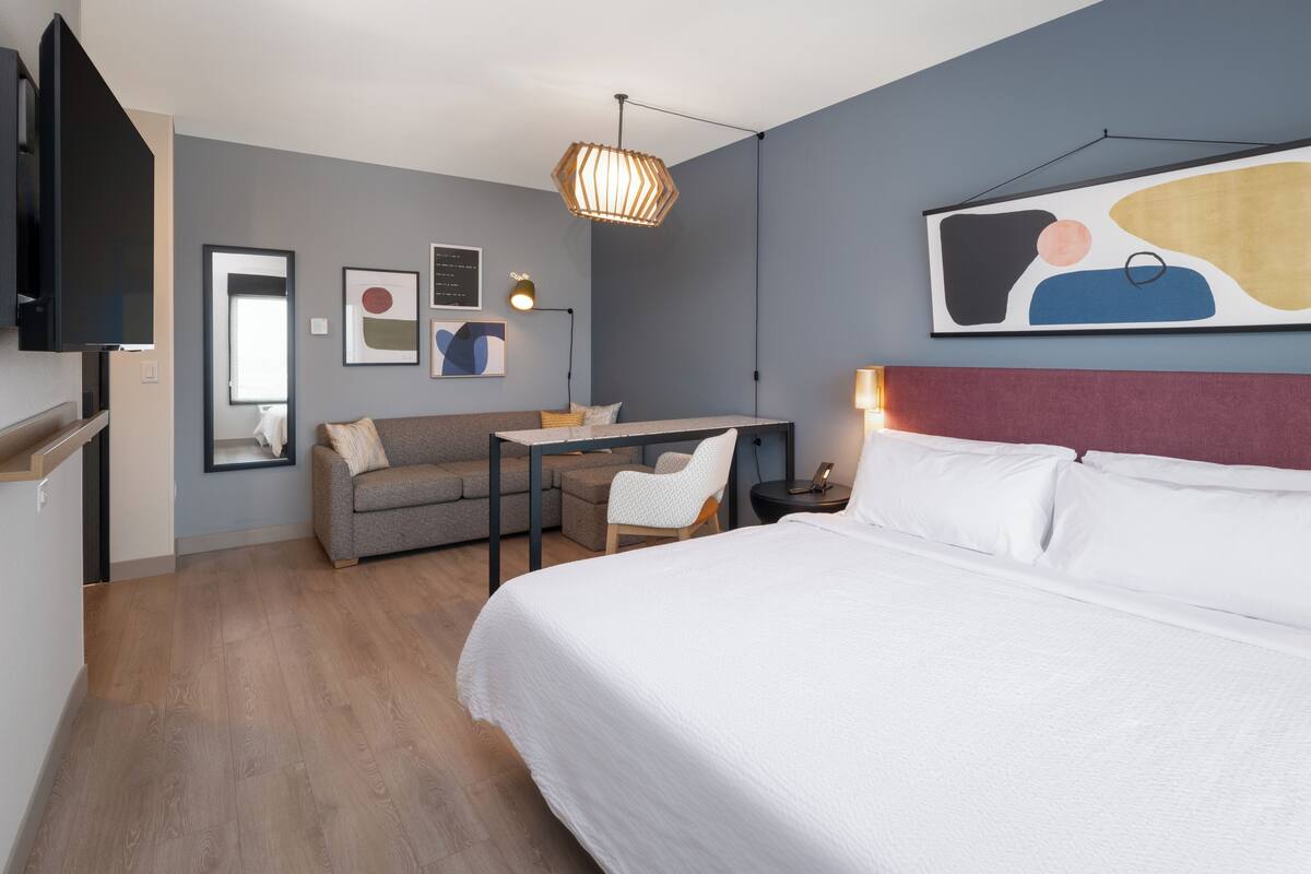 A modern hotel room with a large bed, a desk, a chair, and a TV.