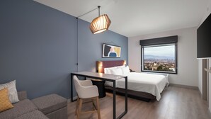 A modern bedroom with a bed, a desk, and a chair. There is a large window with a city view. - Atwell Suites Henderson by IHG (Henderson)