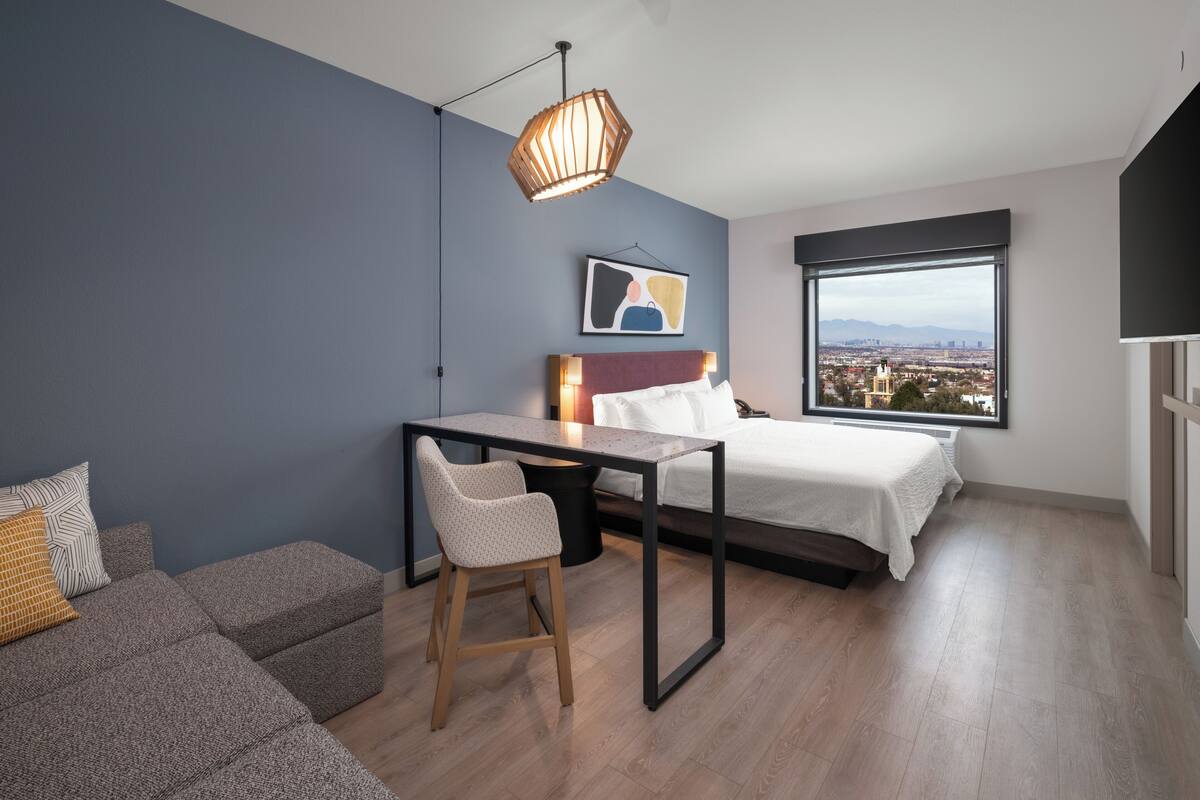 A modern bedroom with a bed, a desk, and a chair. There is a large window with a city view.