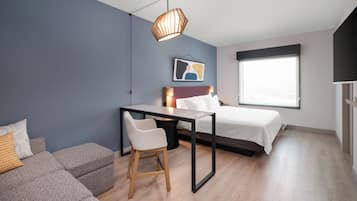 A modern bedroom with a bed, a desk, a chair, a hanging light fixture, and a window.