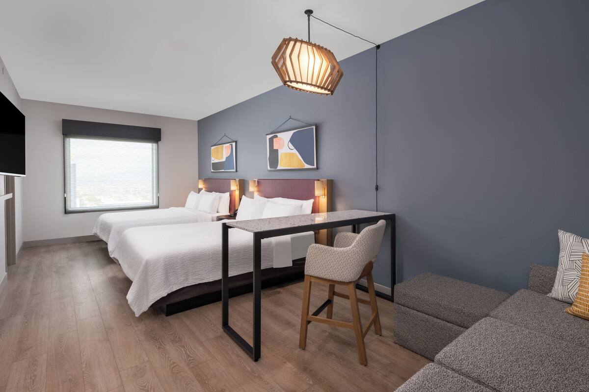 A modern hotel room with a bed, a desk, and a chair. There are two paintings on the wall, a window with blinds, and a ceiling light fixture.