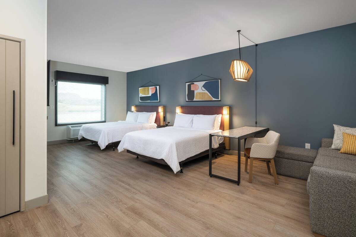 A modern hotel room with two beds, a desk, a chair, and a sofa.