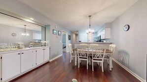 A spacious dining area with a long table and chairs, a kitchen with white cabinets, and a chandelier. - True Blue Golf Getaway 2 Bedroom (Pawleys Island)