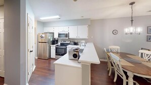 A modern kitchen with white cabinets, a built-in oven, microwave, and a coffee maker. There is a dining area with a wooden table and white chairs. - True Blue Golf Getaway 2 Bedroom (Pawleys Island)