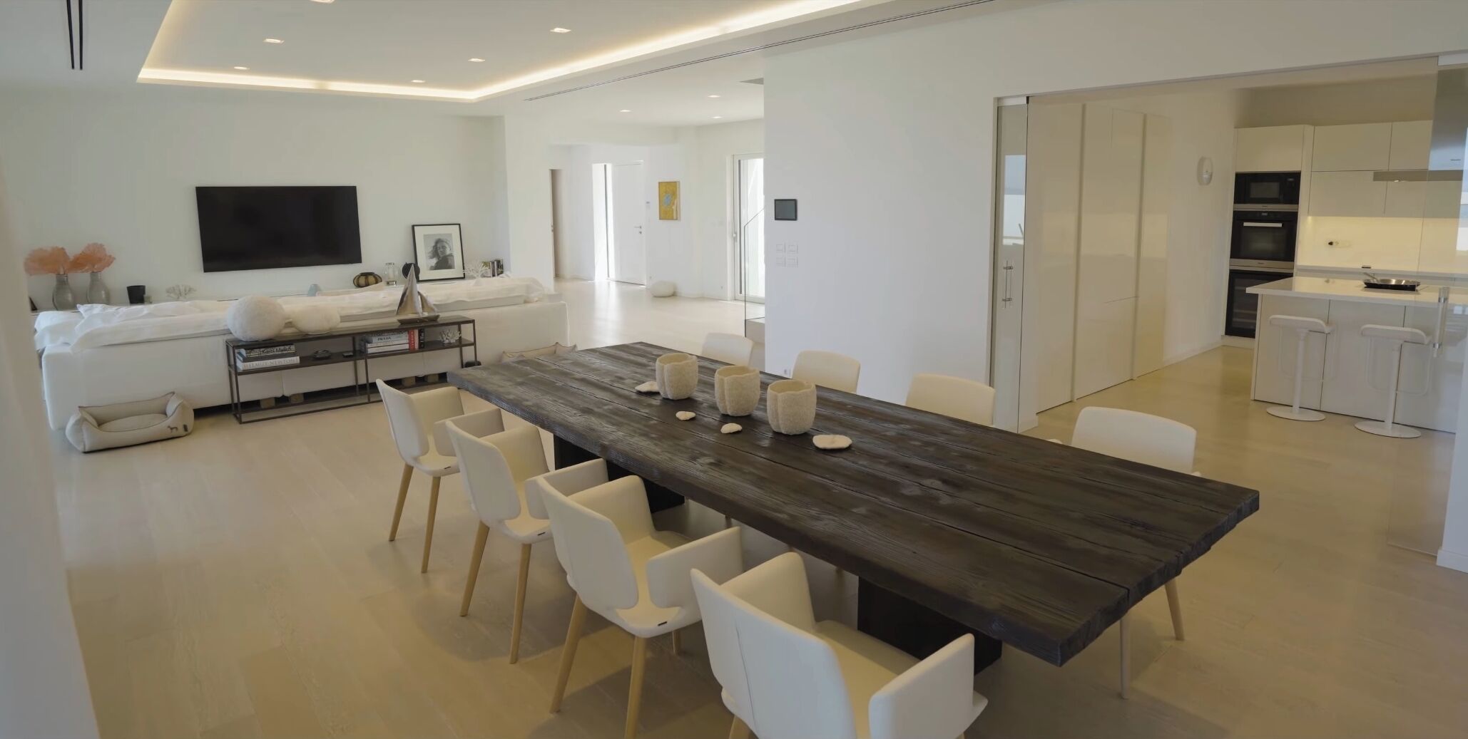 Un salon moderne avec une grande table à manger en bois, des chaises blanches, un canapé et un téléviseur à écran plat.