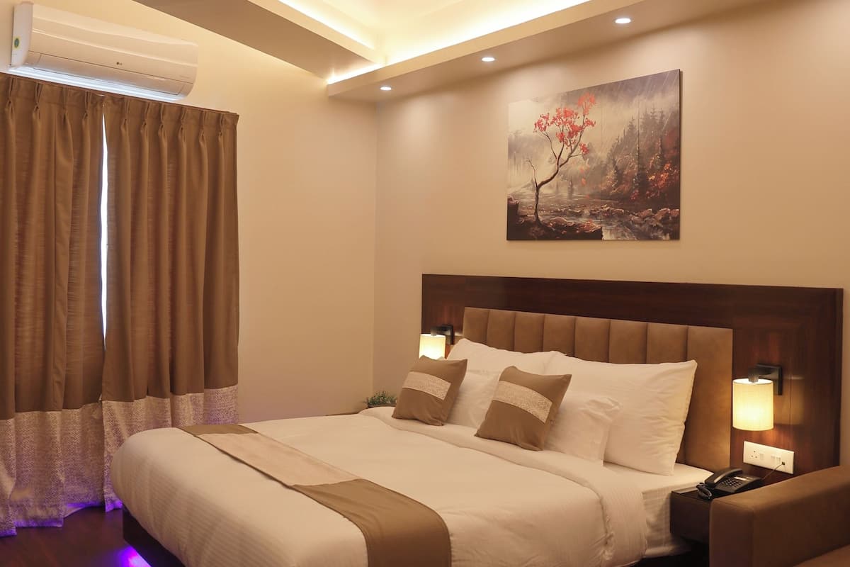 A modern hotel room with a large bed, recessed ceiling lighting, a painting on the wall, and a small table with a lamp.