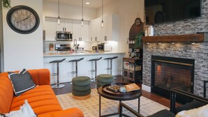 En moderne stue med sofa, skamler, sofabord, pejs og et køkken i baggrunden.