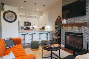 Un salon moderne avec un canapé, des poufs, une table basse, une cheminée et une cuisine en arrière-plan.