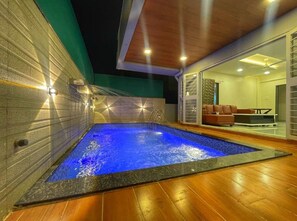 An indoor swimming pool with a blue-lit water feature, surrounded by a wooden deck and a modern interior with a sofa and a glass door leading to a bright room. - Sunshine Villa Igatpuri (Igatpuri)