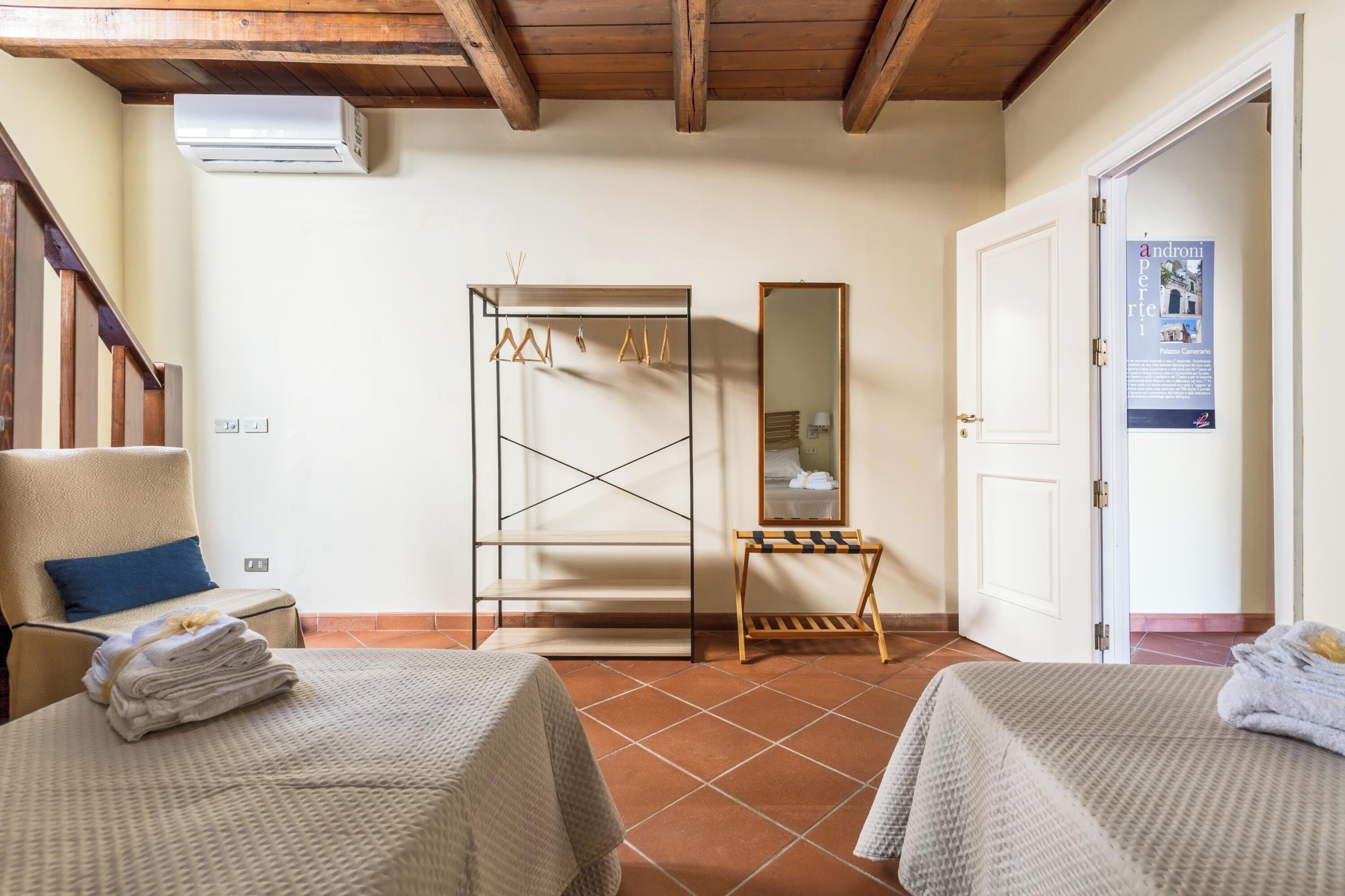 Quarto de hotel com uma cama, uma cadeira, um guarda-roupa de madeira, um espelho e uma porta.