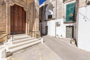 A narrow street with a large wooden door and steps leading to a building with a white facade and green shutters. - 4060 La Loggia House by Barbarhouse (Manduria)
