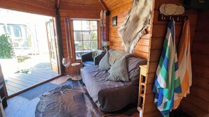 A log cabin interior with a wooden ceiling, a sofa with pillows, a mounted animal hide, and a window with a view of the outside.