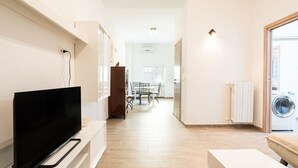 A modern living room with a flat-screen TV on a stand, a dining area with a table and chairs, and a laundry area with a washing machine. - Venus Apartment by Wonderful Italy (Bari)