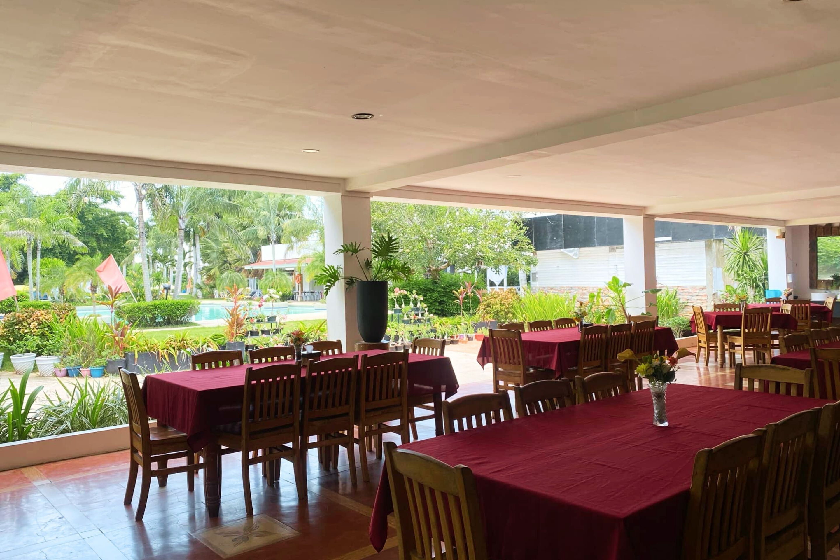 A restaurant with multiple tables covered in red tablecloths, wooden chairs, and potted plants.