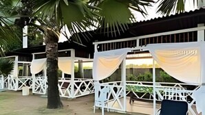 Un espacio al aire libre techado con estructuras de celosía blanca, una silla de descanso y una palmera.