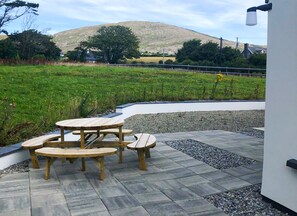 A wooden picnic table set on a stone patio with a view of a grassy field and a hill in the background. - Bell View Holiday Home Sheeps Head (Kilcrohane)