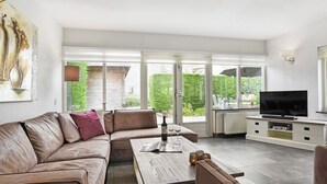 A modern living room with a leather sofa, a coffee table, a flat-screen TV, and a large window with blinds. - Bungalow for 4 persons with enclosed garden (Brouwershaven)