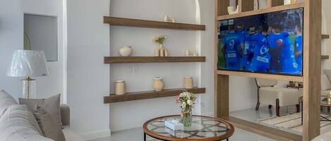 A modern living room with a sofa, coffee table, and TV. There are shelves with decorative items and a vase with flowers.